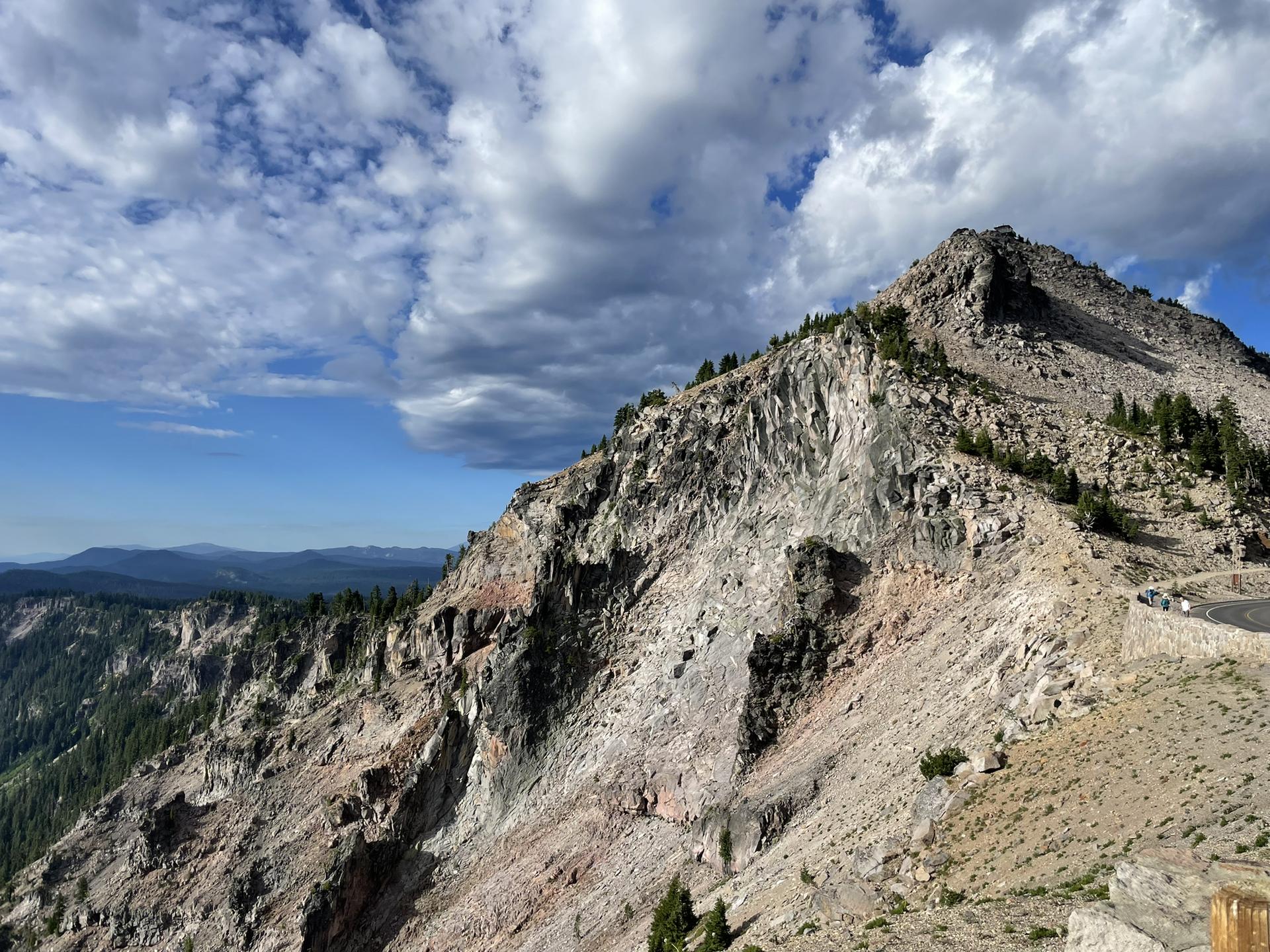 Steep, rocky mountain slope with sparse trees under a cloudy sky.