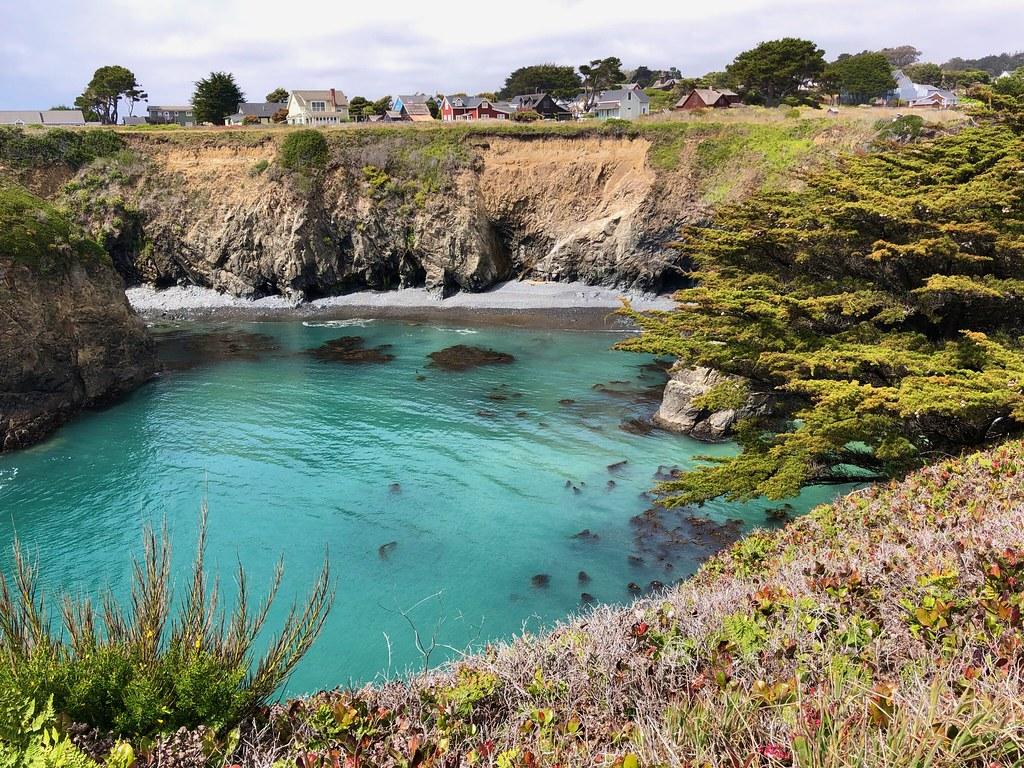 Turquoise cove with rocky cliffs and distant houses under an overcast sky.