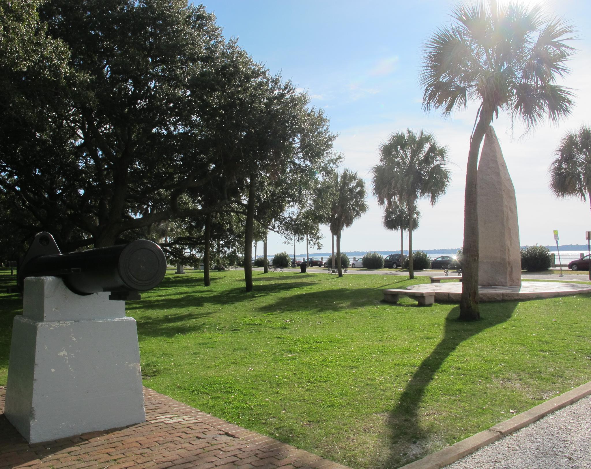 A sunny park with a cannon, palm trees, and water in the distance.