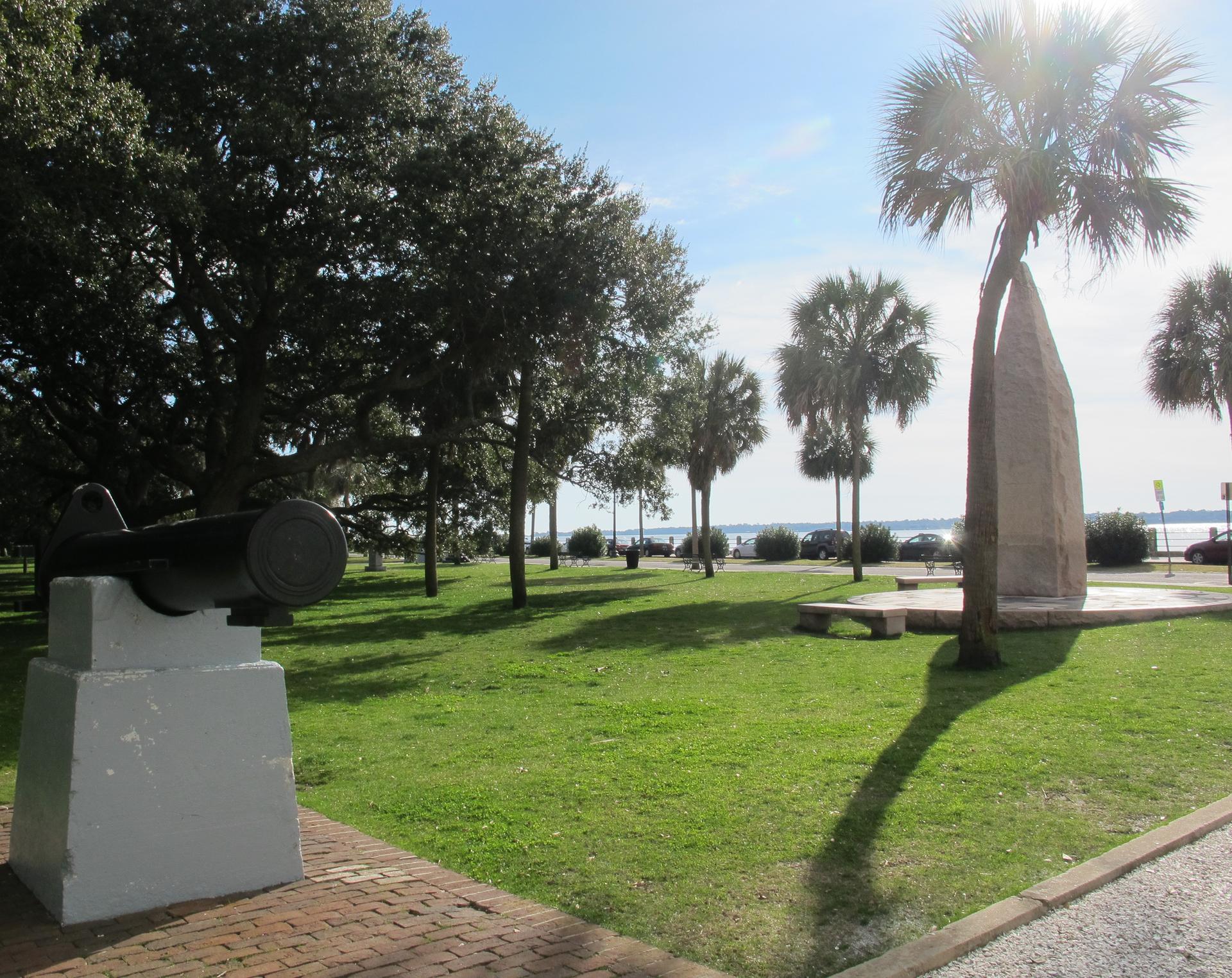 A sunny park with a cannon, palm trees, and water in the distance.