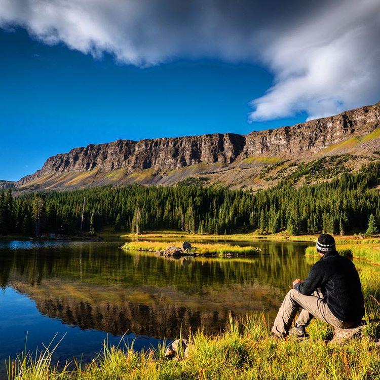 Hiker sits by a calm lake reflecting mountains and forest under a bright sky.
