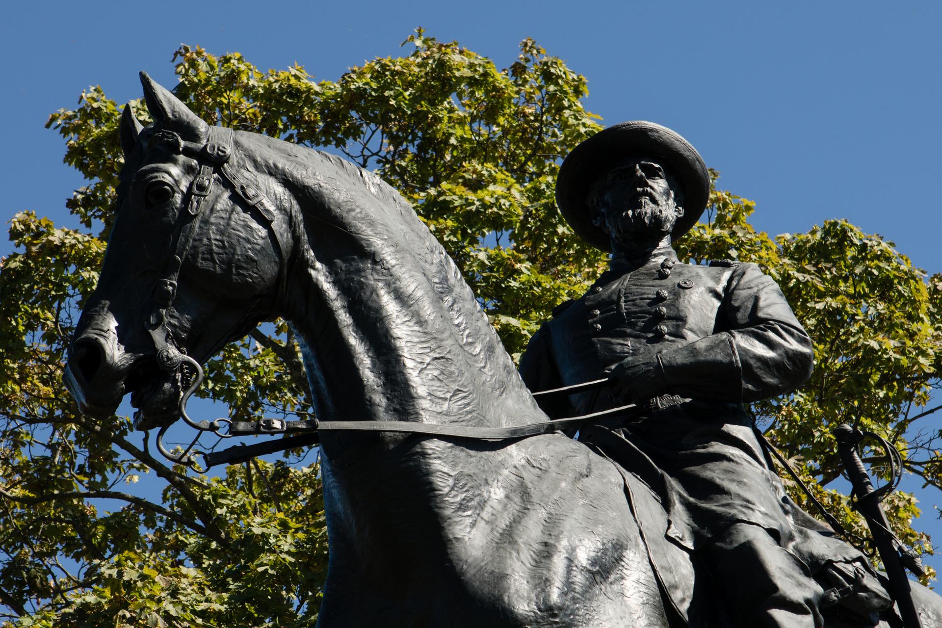 Bronze equestrian statue of a man on horseback against a blue sky and green trees.
