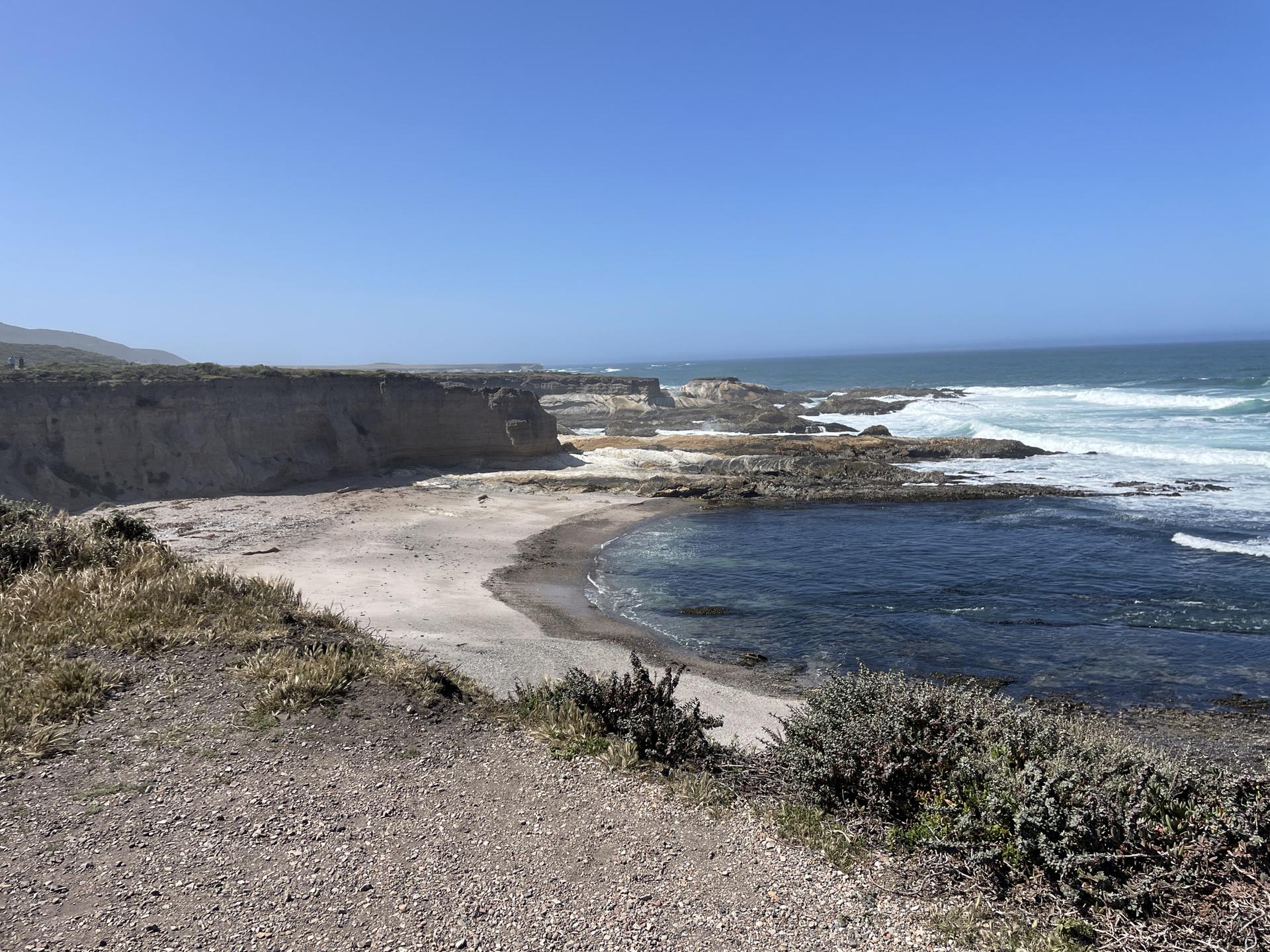 Sunny coastal view with rugged cliffs, a sandy cove, and ocean waves.