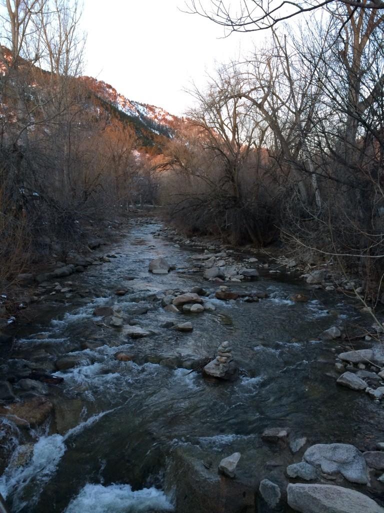 A rocky river flows through bare trees with mountains in sunset light.