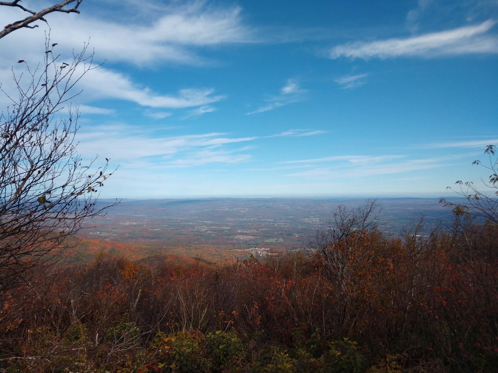 Expansive valley view from a mountain, with autumn trees and blue sky.