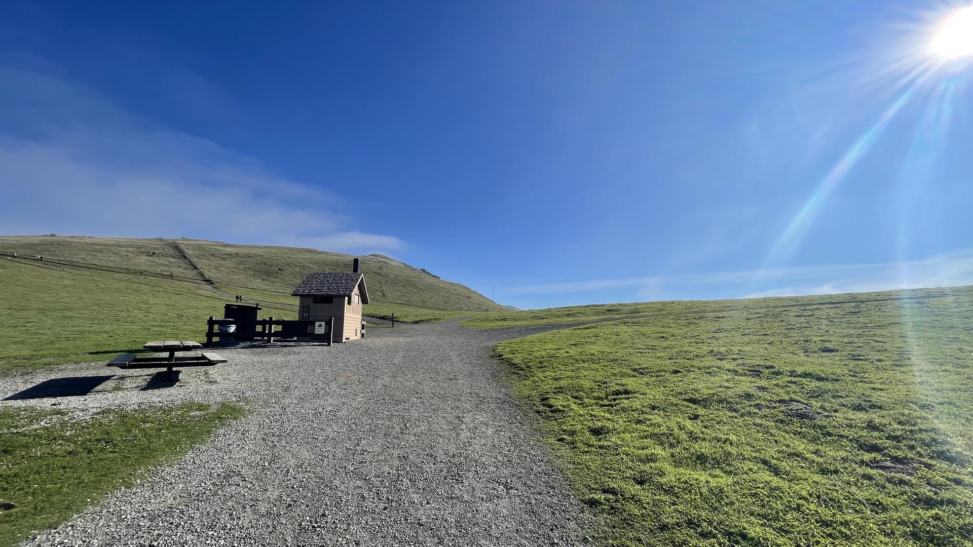 A small wooden building and picnic table next to a gravel path on a sunny green hill.