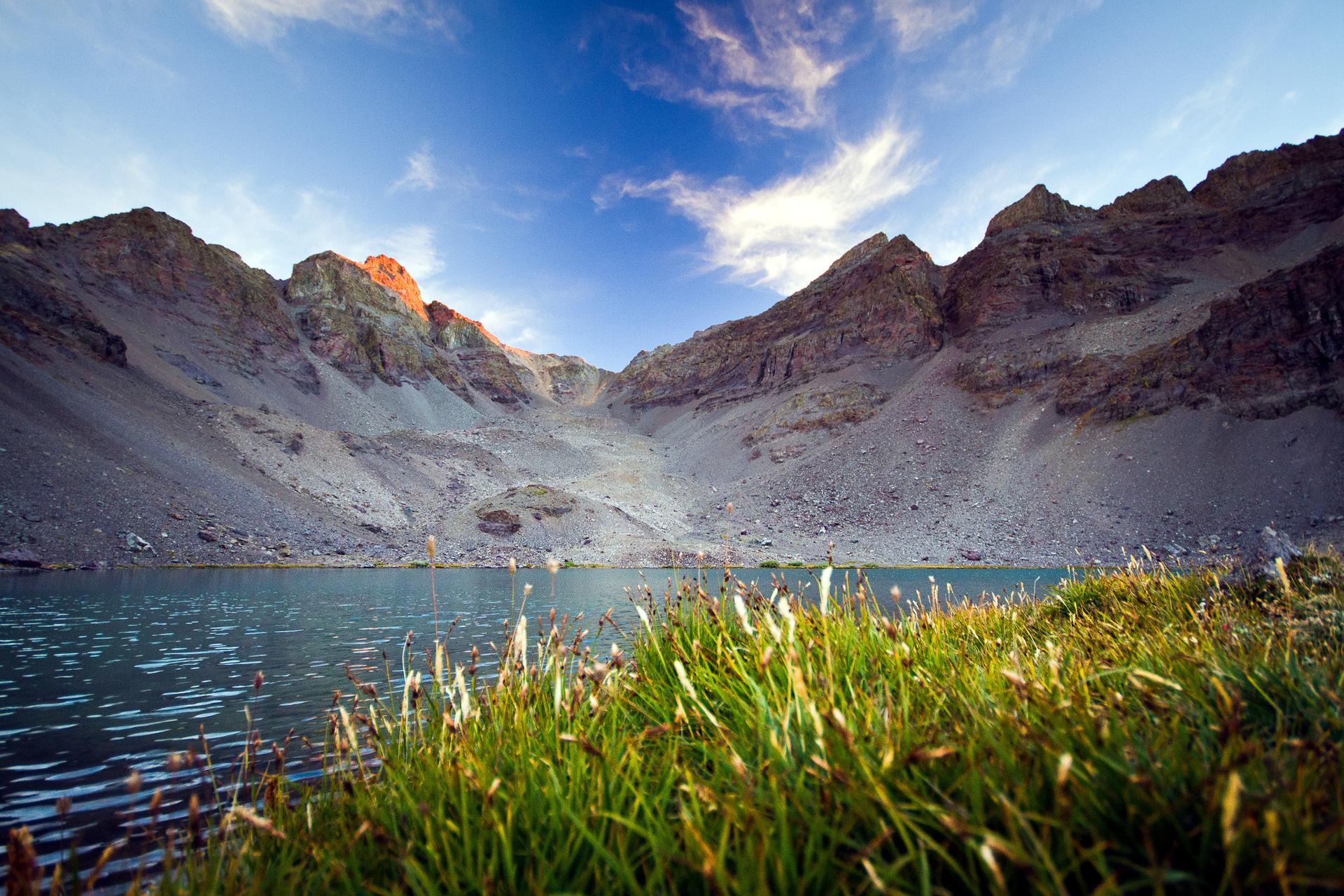 Golden light illuminates rocky mountains overlooking a serene lake with foreground grass.