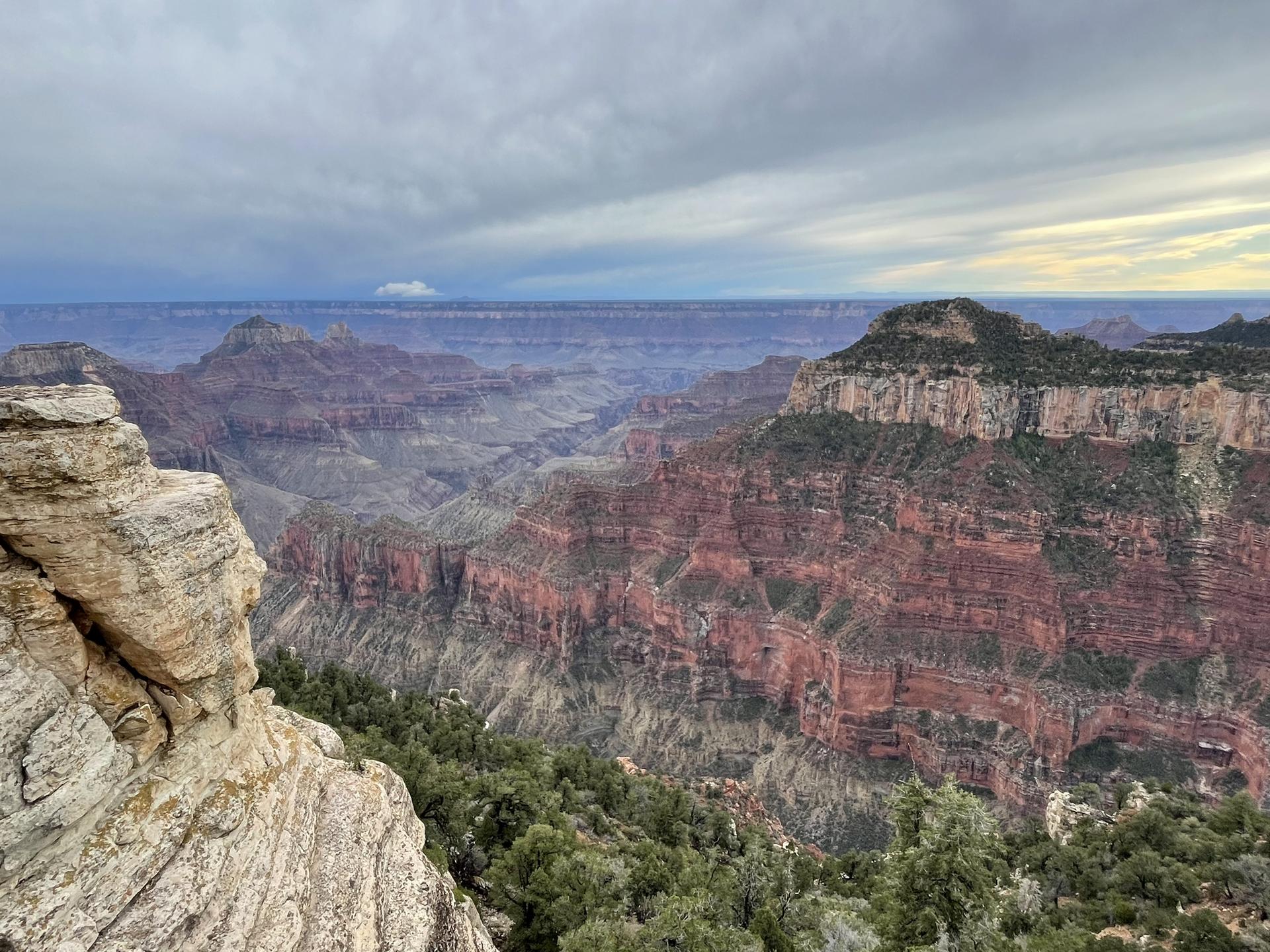 Vast canyon landscape under a dramatic, cloudy sky, with rocky foreground.