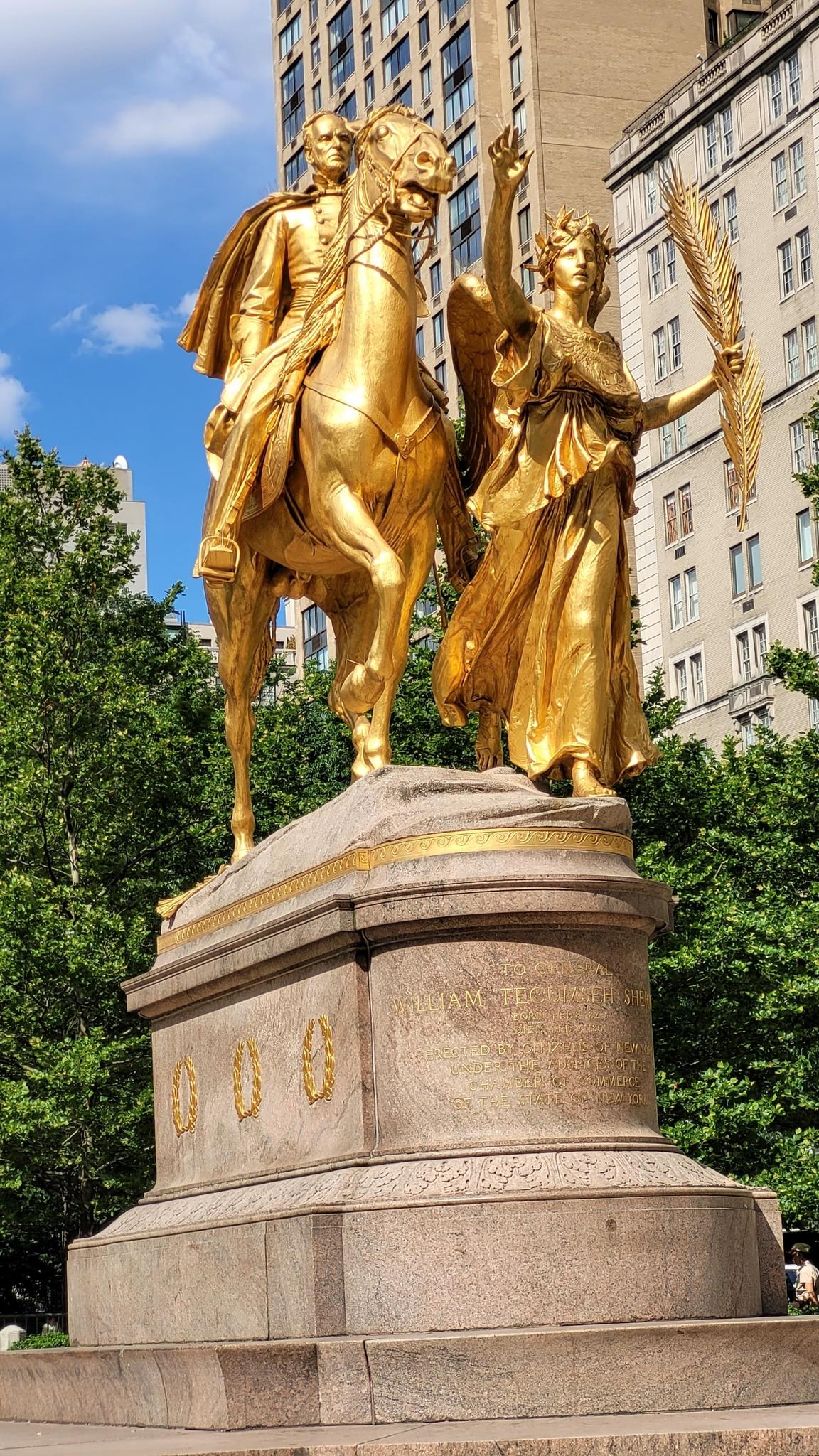 Golden equestrian statue with an allegorical figure, surrounded by trees.