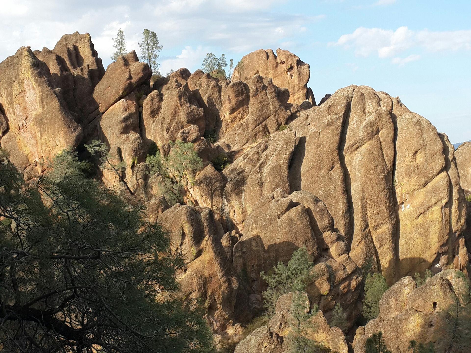Jagged, brown rock formations with sparse green trees under a partly cloudy sky.