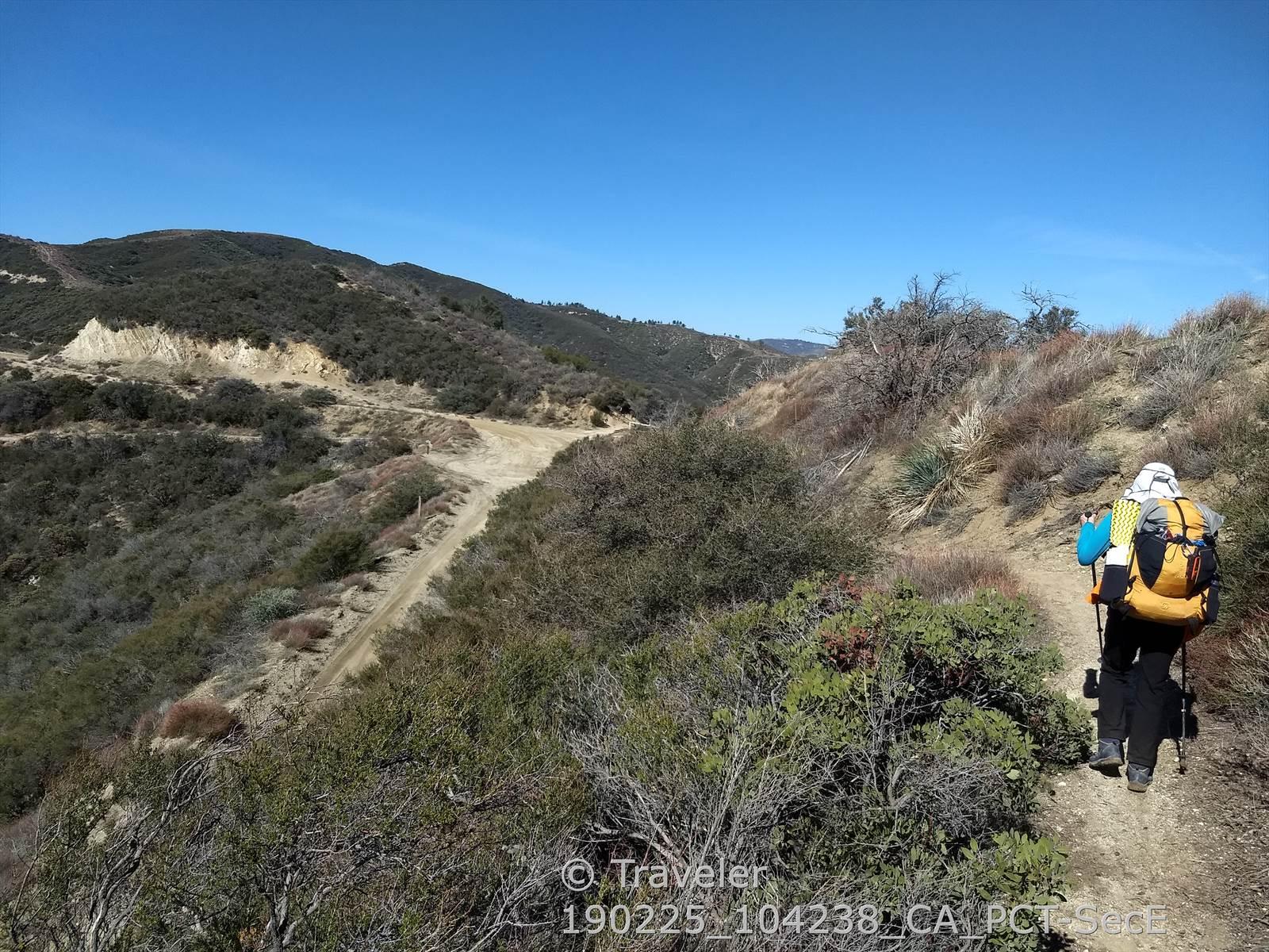 Hiker with a large yellow backpack walks on a dirt trail through scrubby hills.