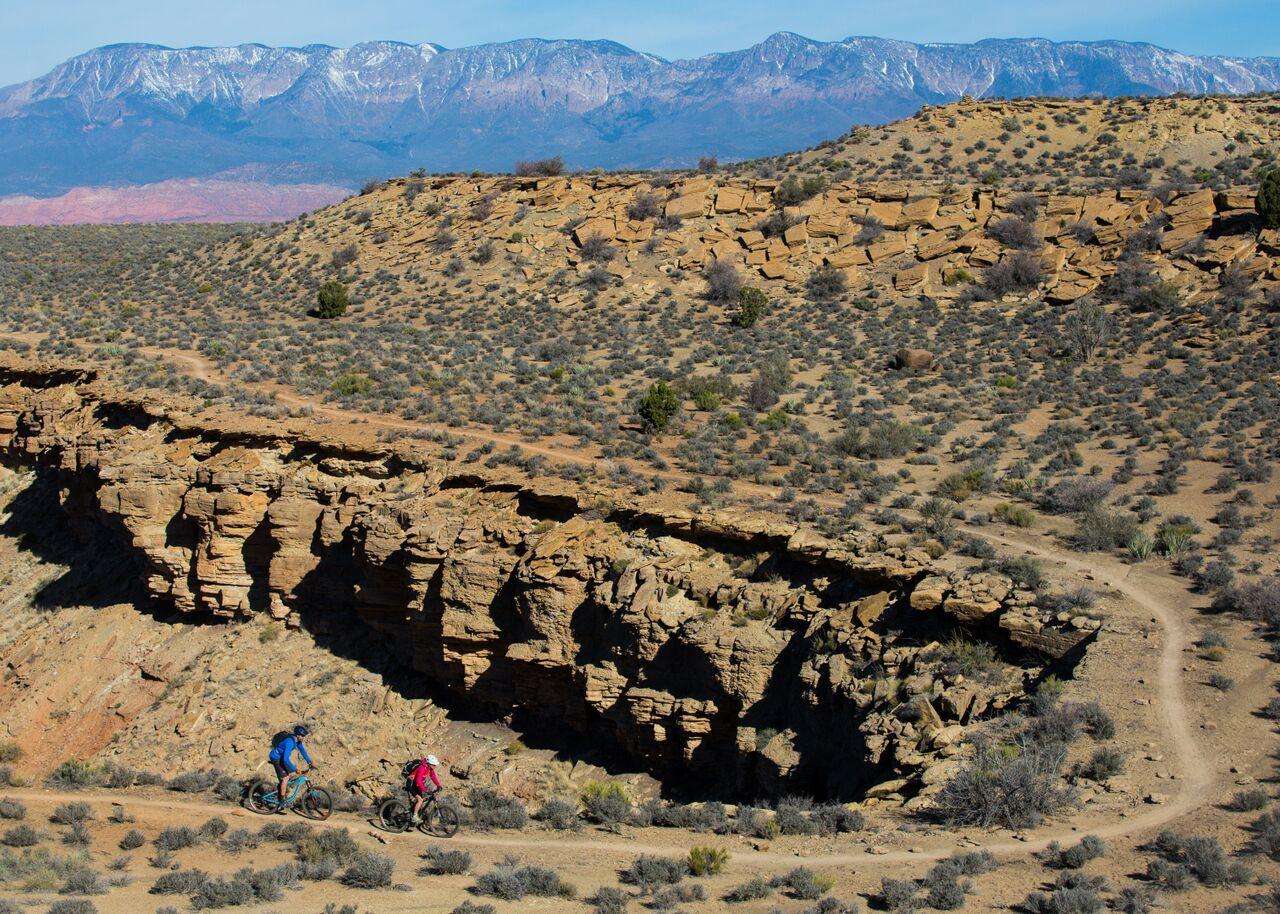 Two mountain bikers on a desert trail with distant snow-capped mountains.