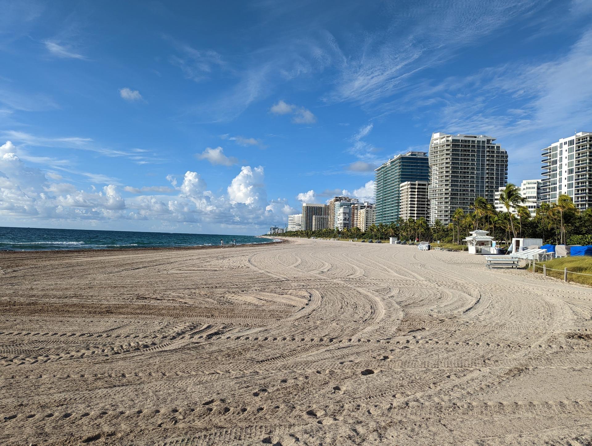 Expansive sandy beach with tire tracks, ocean, and city skyline.