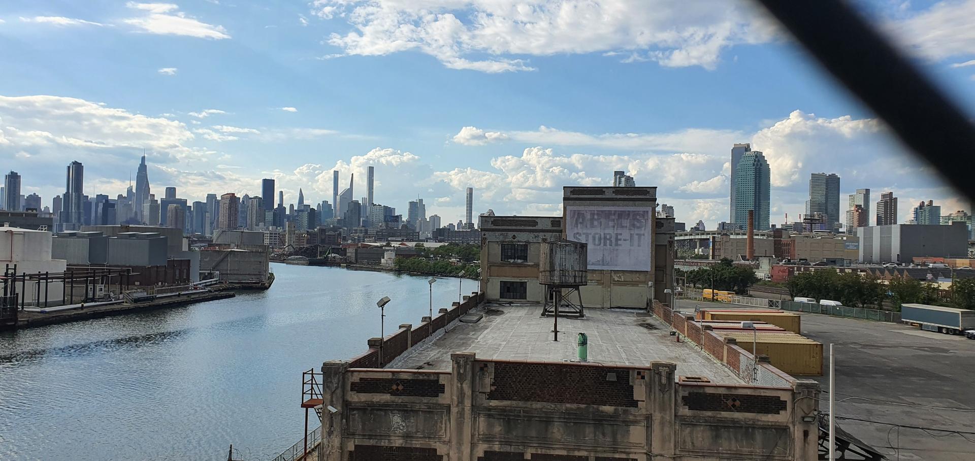 Urban waterfront with river, industrial buildings, and distant city skyline.
