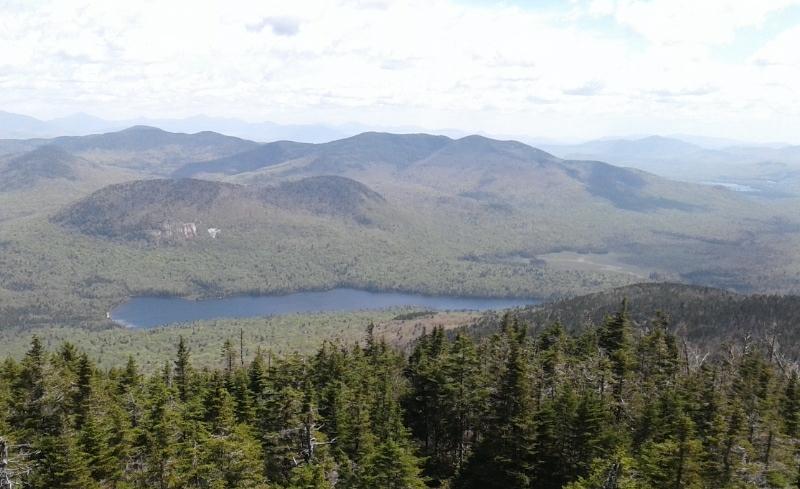 Expansive mountain vista with a lake, lush green forests, and cloudy skies.
