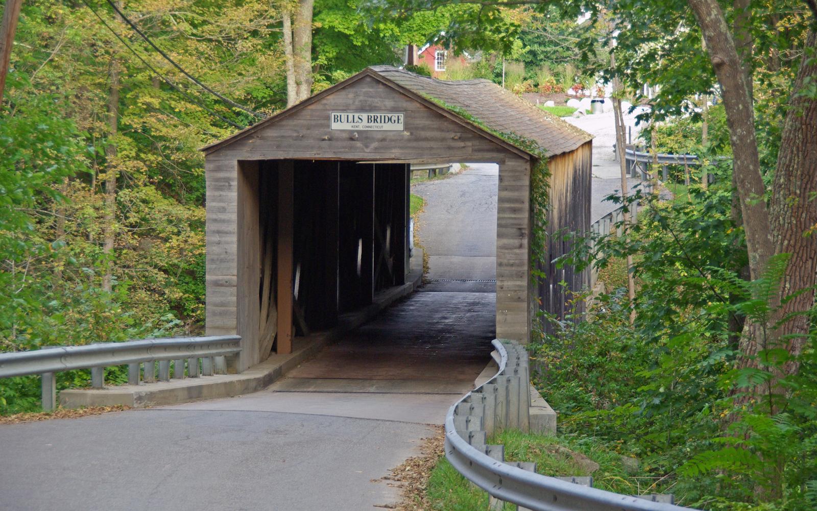 Rustic wooden covered bridge over a road, surrounded by green trees.