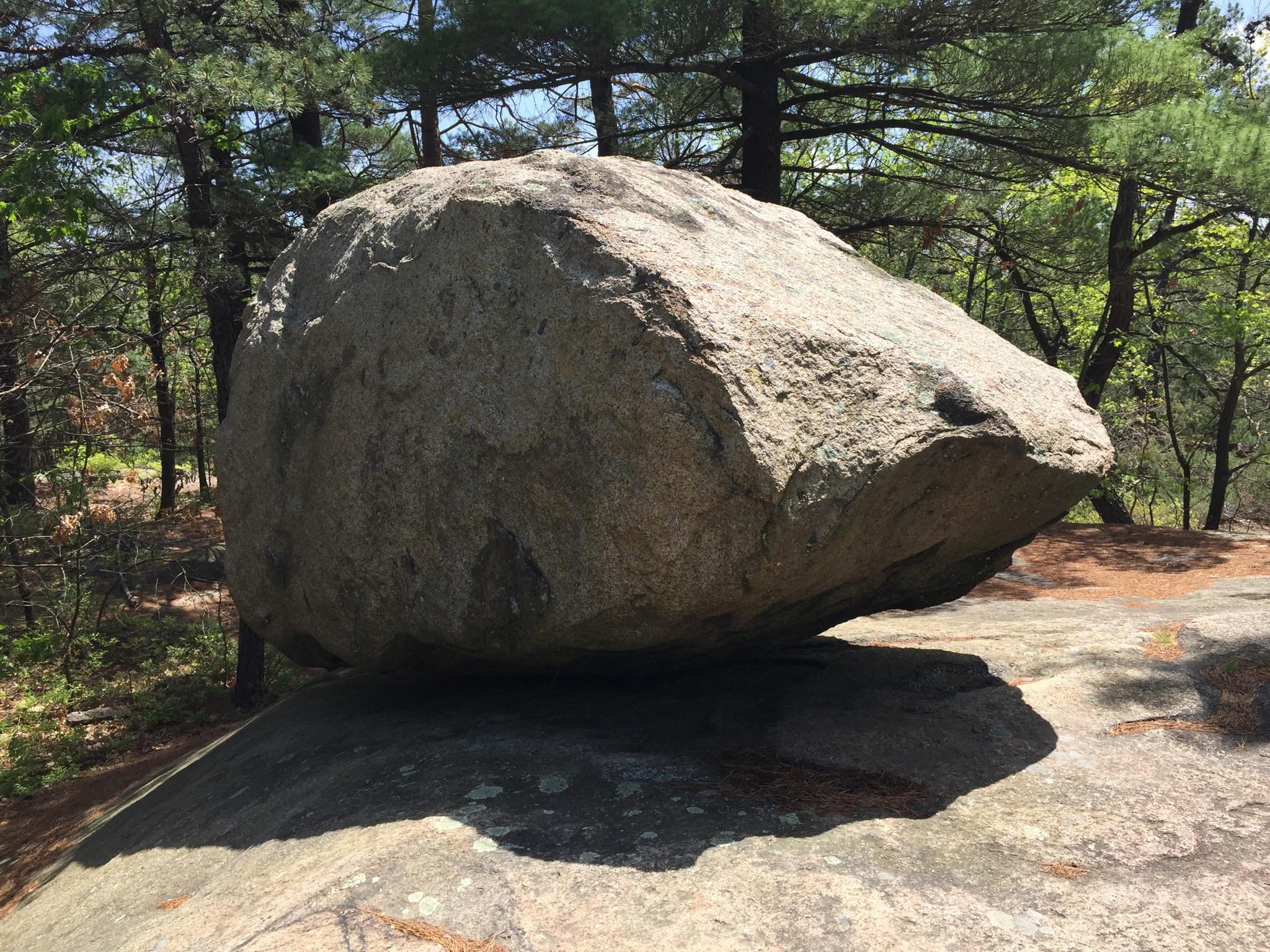 Large, weathered boulder resting on a rock slab in a sunlit forest.