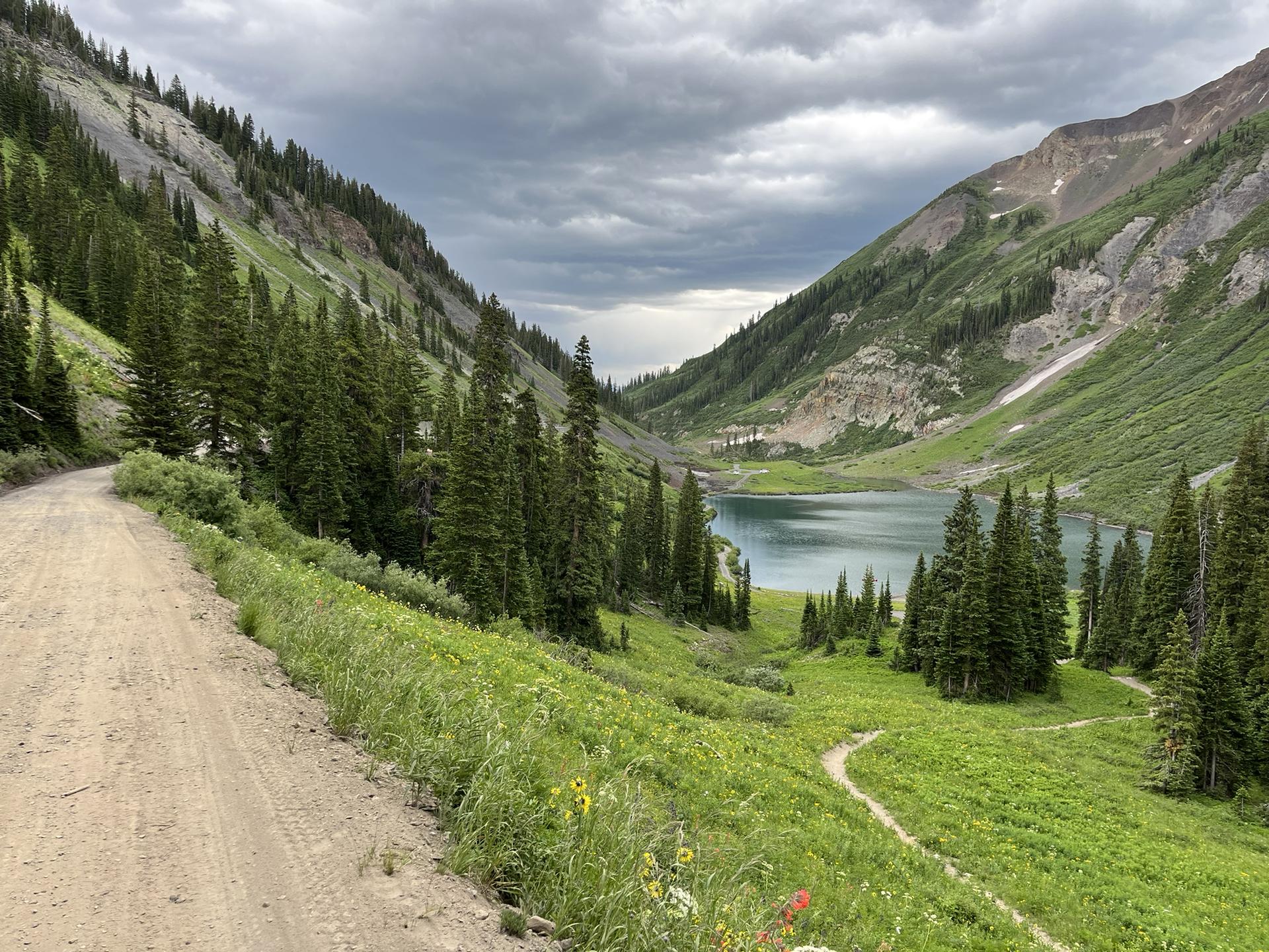 Dirt road overlooking a mountain lake surrounded by green slopes under cloudy skies.