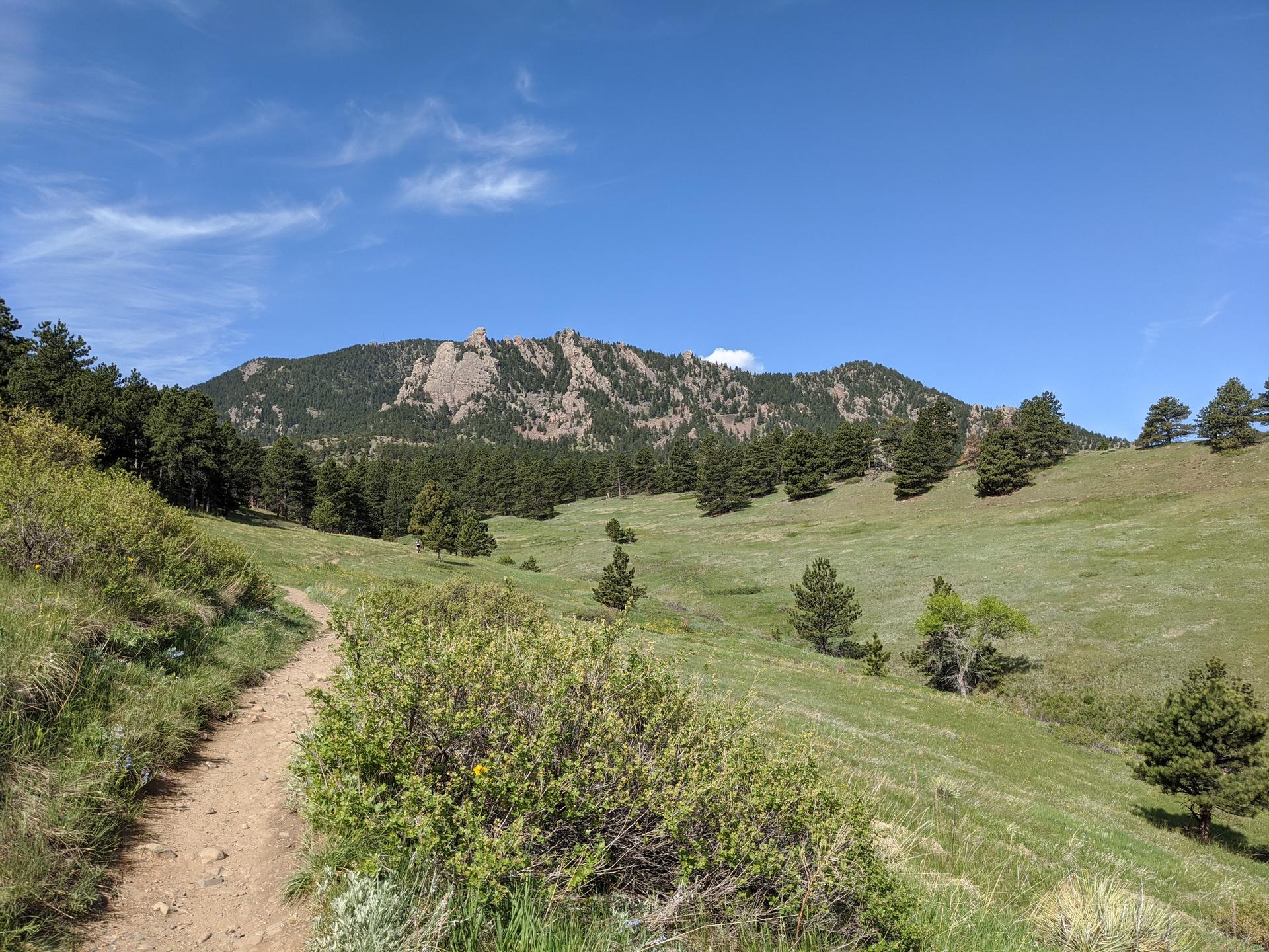 Dirt path winds through green hills towards a large, rocky mountain.