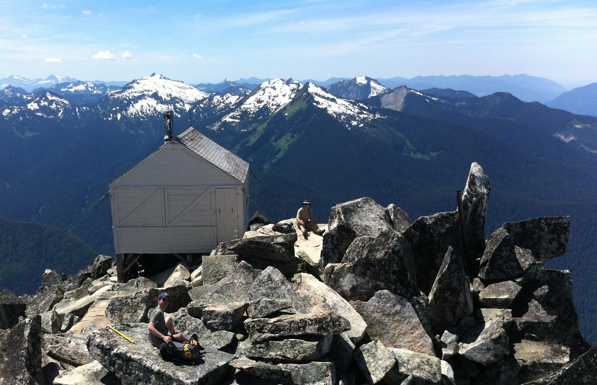 Two people near a small cabin on a rocky peak, snowy mountains beyond.