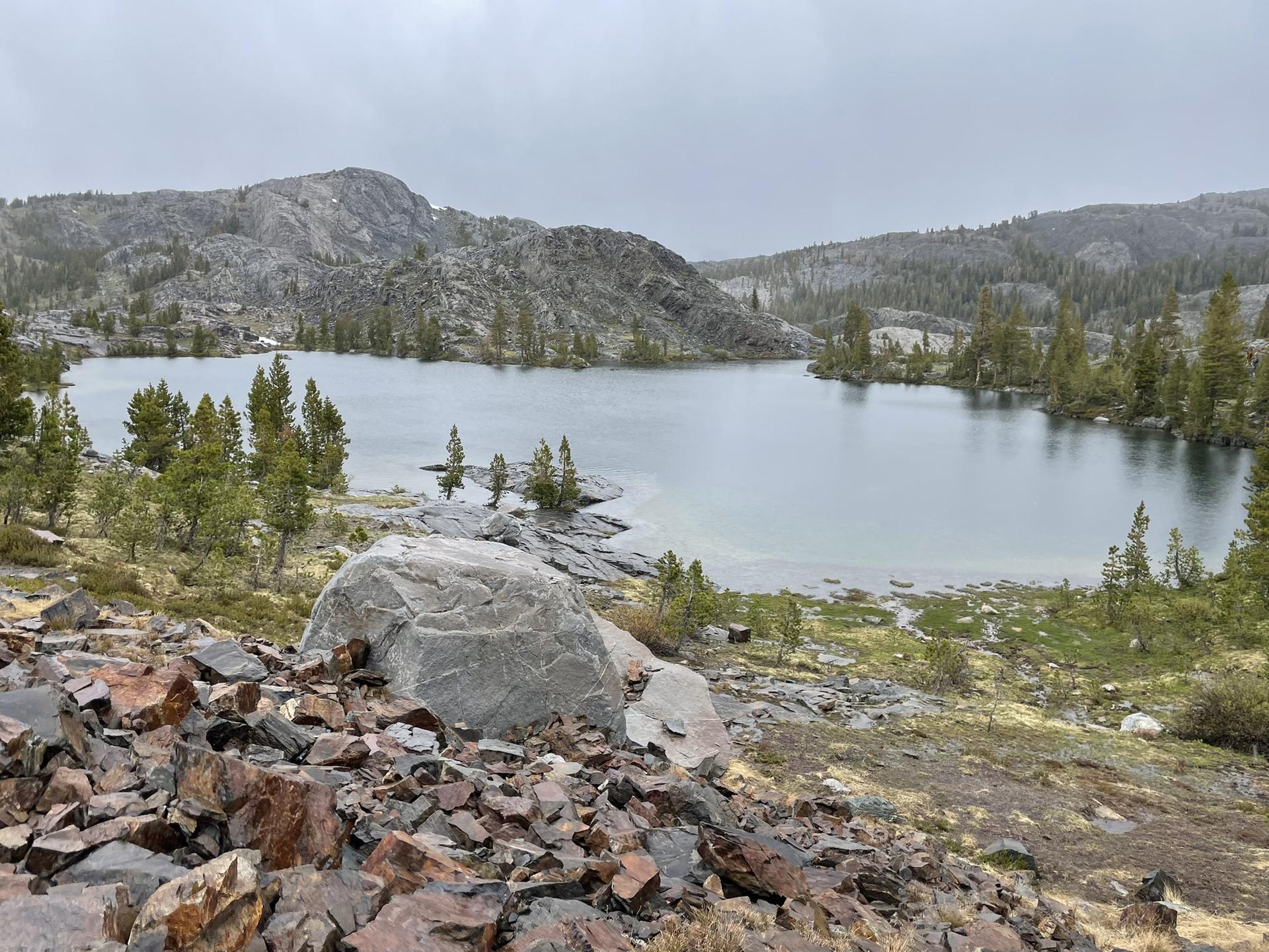 Overcast view of a calm lake surrounded by rocky hills and sparse pine trees.