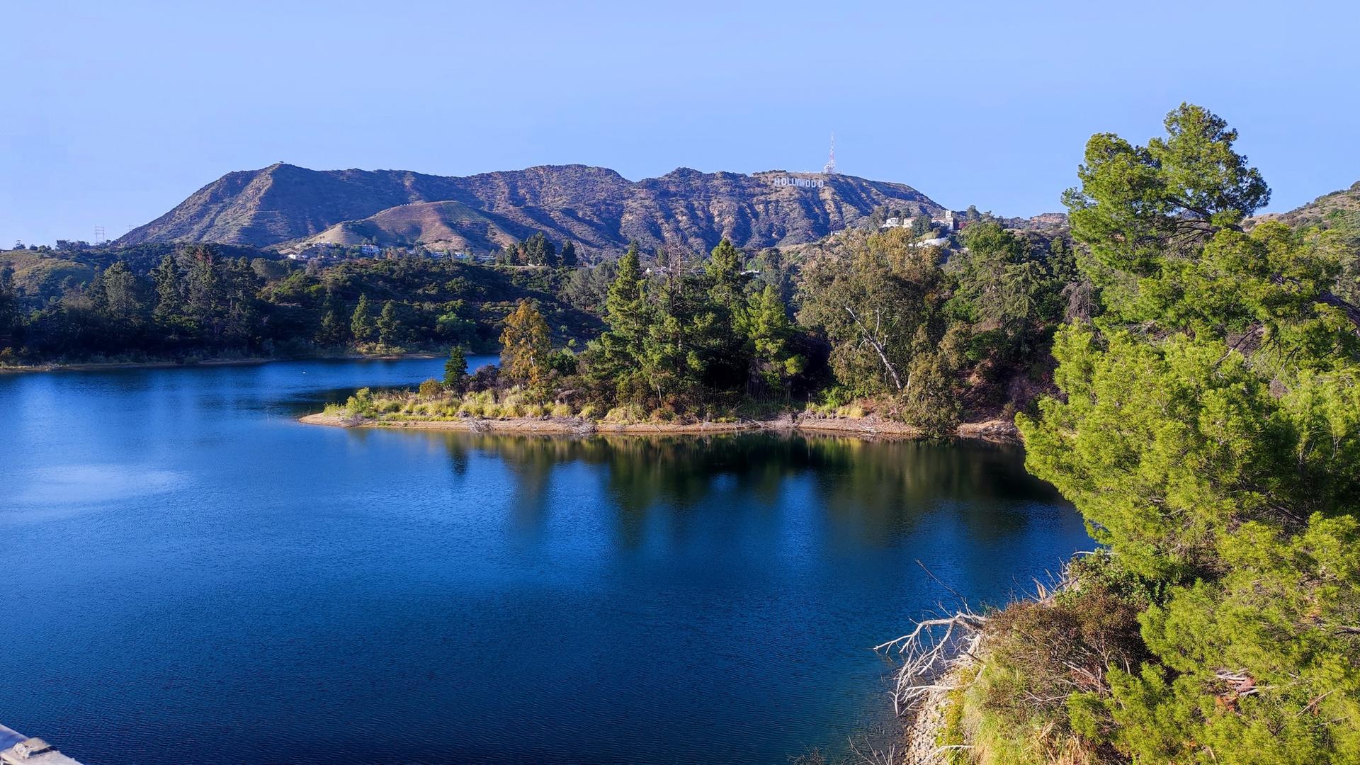 Blue lake with green trees and a distant mountain range under clear sky.
