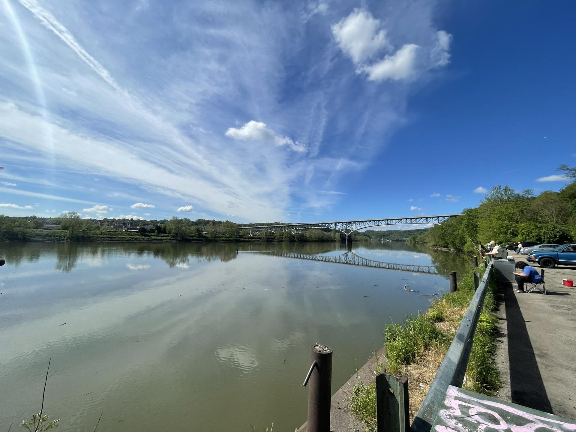Wide river under a bright sky, distant bridge, person fishing on bank.