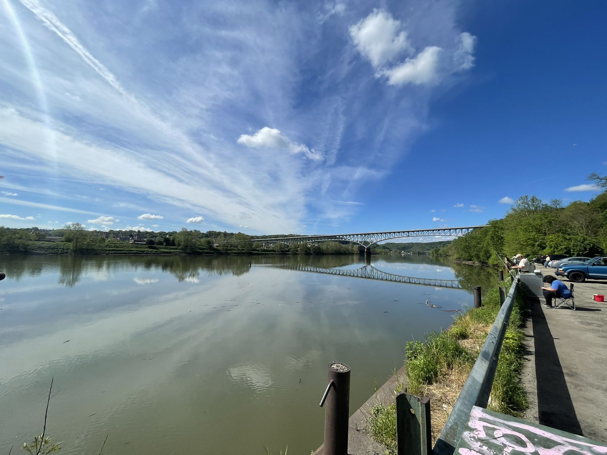 Wide river under a bright sky, distant bridge, person fishing on bank. - Duck Hollow Trail
