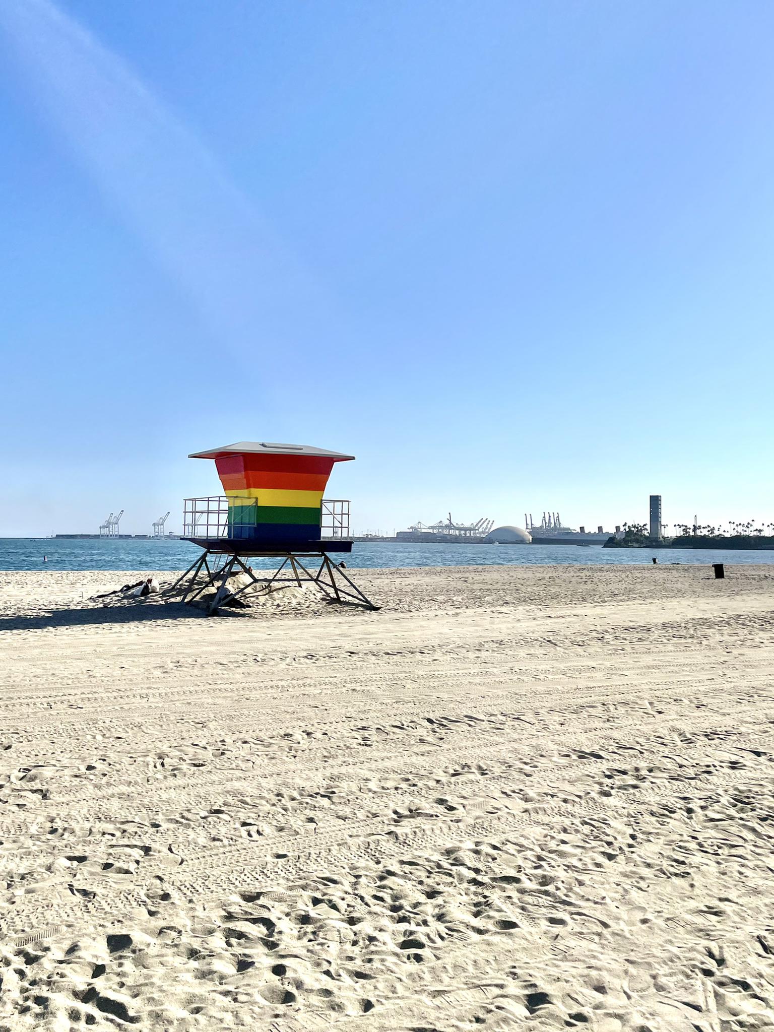 Rainbow-colored lifeguard stand on a wide sandy beach under a clear blue sky.
