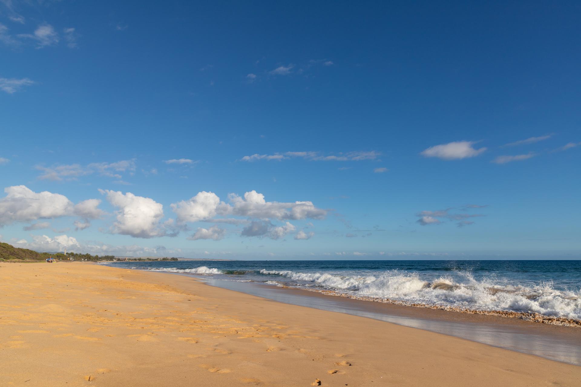 Sunny beach with golden sand, blue ocean waves, and scattered clouds.