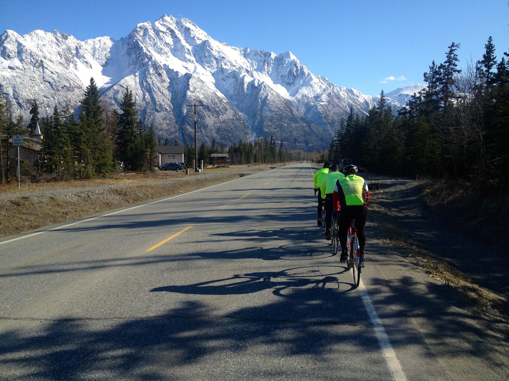 Three cyclists ride on a road with snow-capped mountains in sunlight.