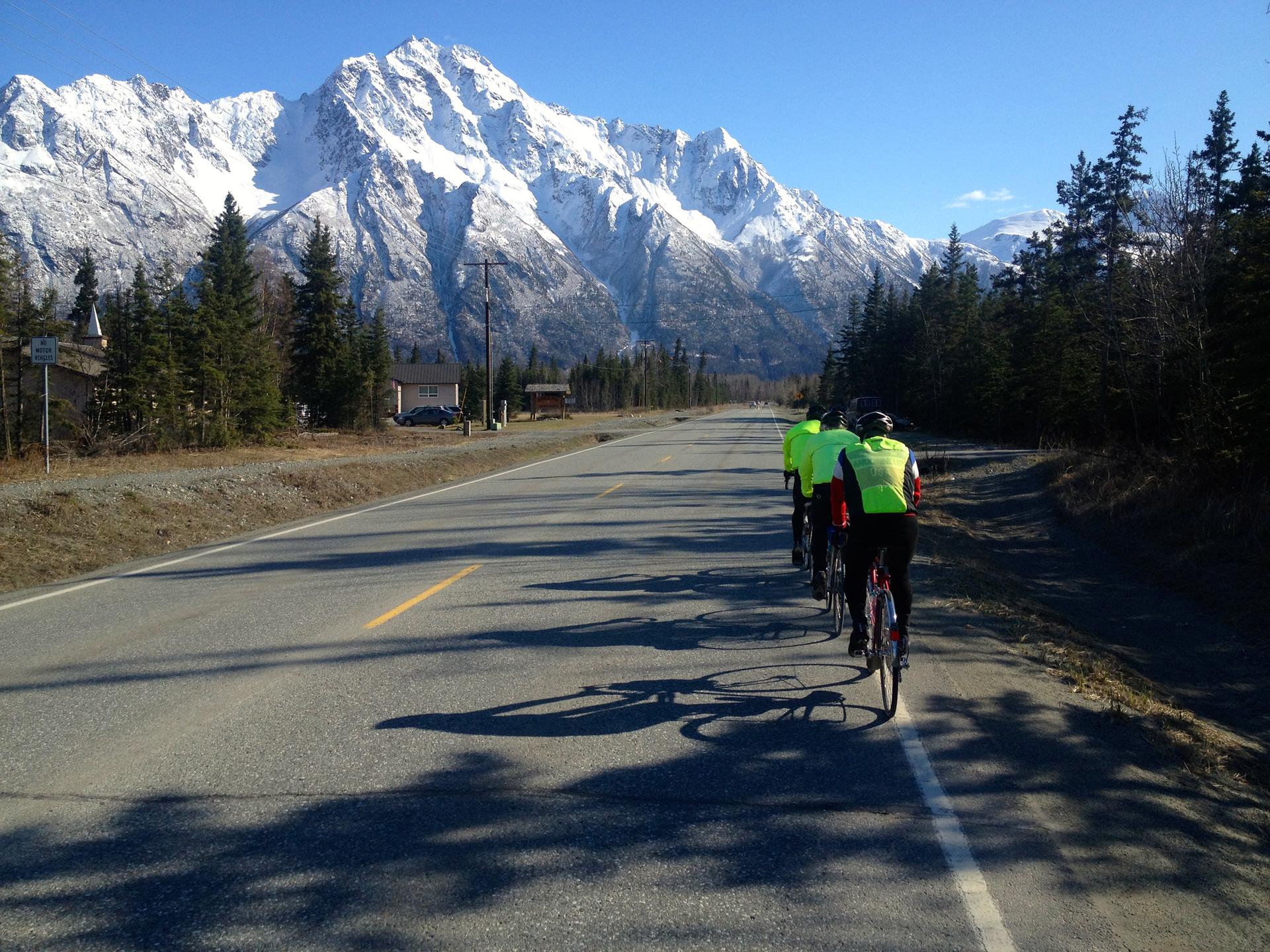 Three cyclists ride on a road with snow-capped mountains in sunlight.
