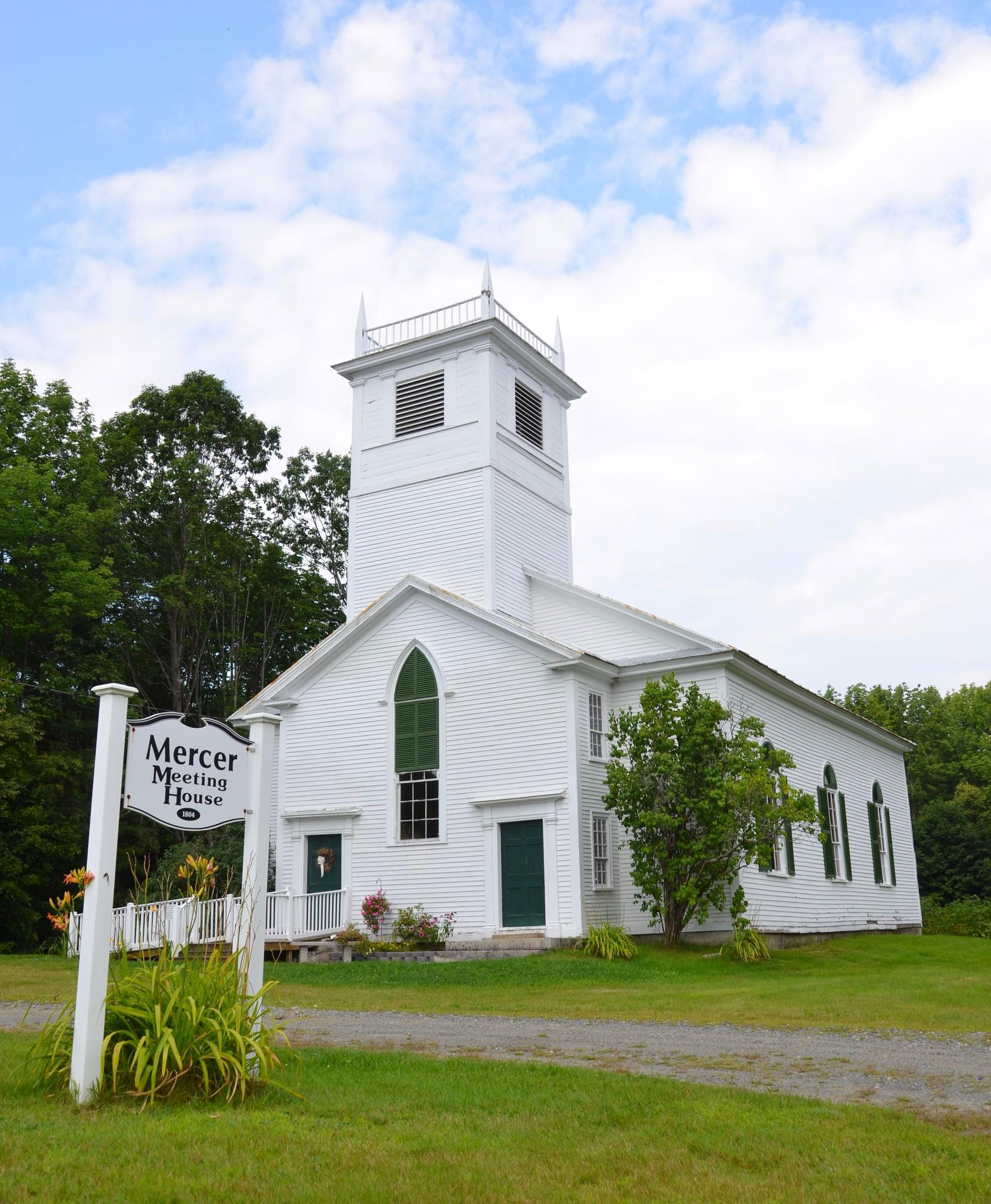 A white church with a tall steeple, green doors, surrounded by trees, bright day.