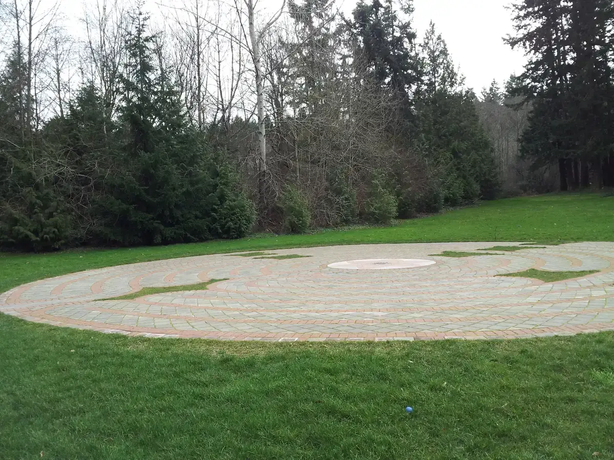 Circular paved labyrinth on green grass surrounded by trees under an overcast sky.