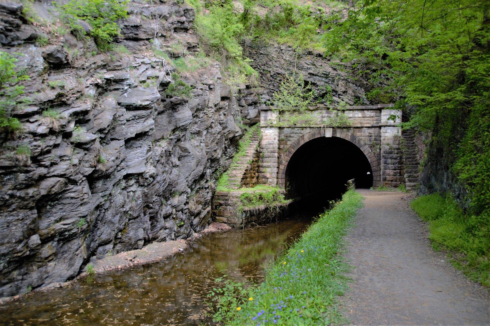 Stone tunnel entrance next to a canal and dirt path, surrounded by rocky cliffs.