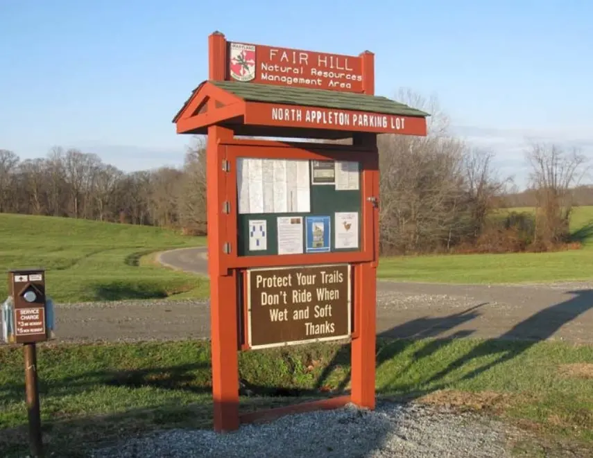 A wooden information kiosk with a brown sign in a sunny, grassy area.