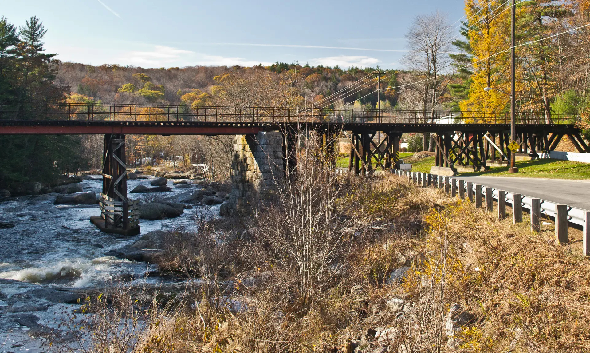 A long bridge crosses a rocky river with autumn trees under bright daylight.