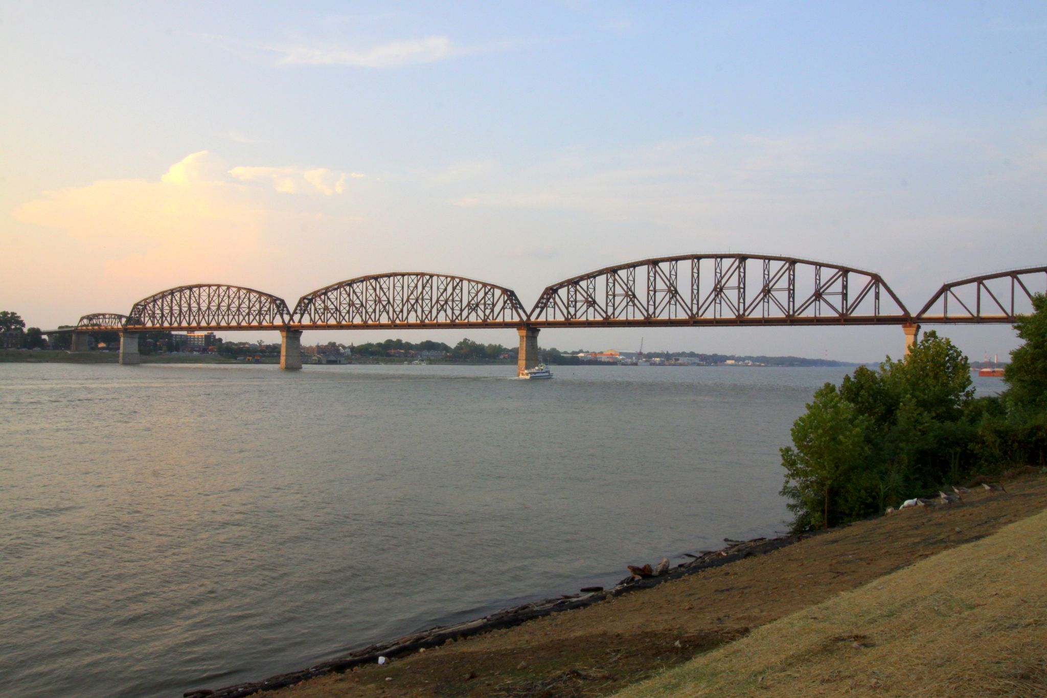 A long truss bridge crosses a wide river with a boat, under soft evening light. - Louisville Loop