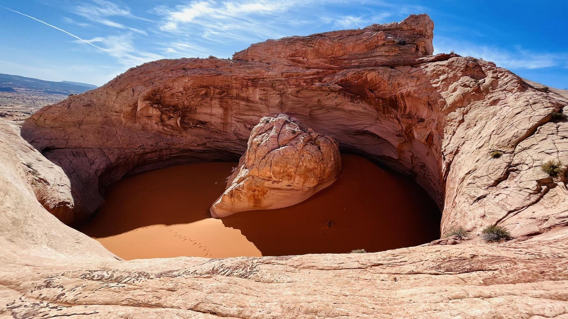 Massive rock formation surrounds a deep, orange sand-filled basin.