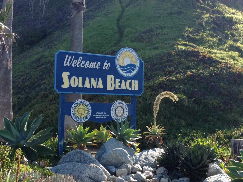 A blue welcome sign with a sun logo, surrounded by green plants.