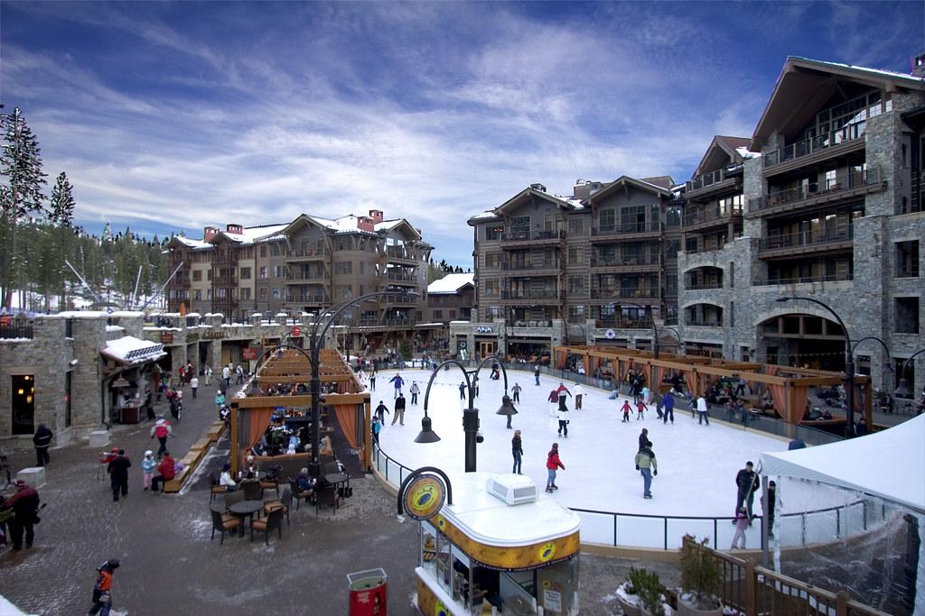 People ice skating on an outdoor rink surrounded by buildings under a bright blue sky.