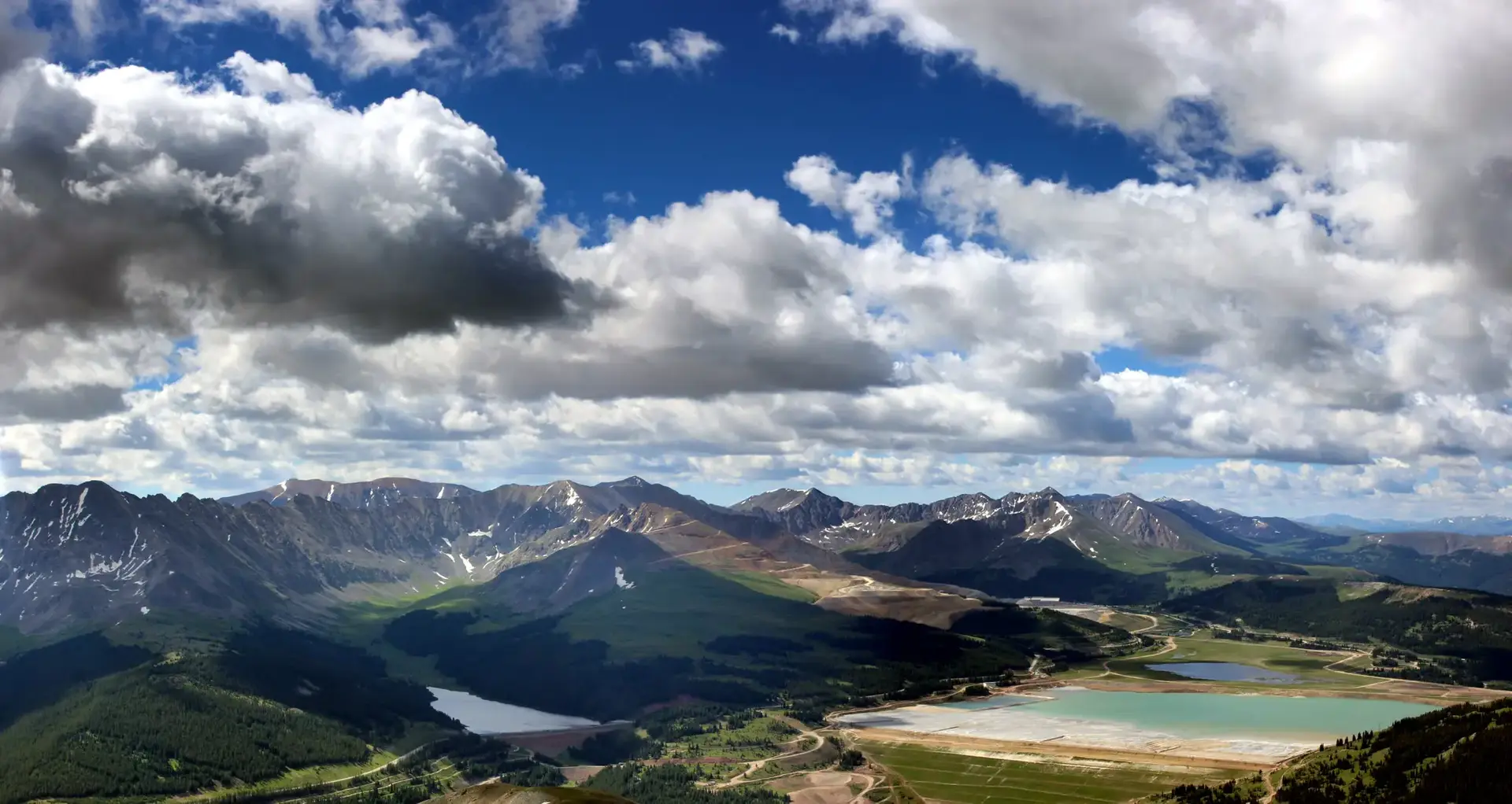 Vast mountain landscape with lakes, roads, and dramatic clouds under bright skies.