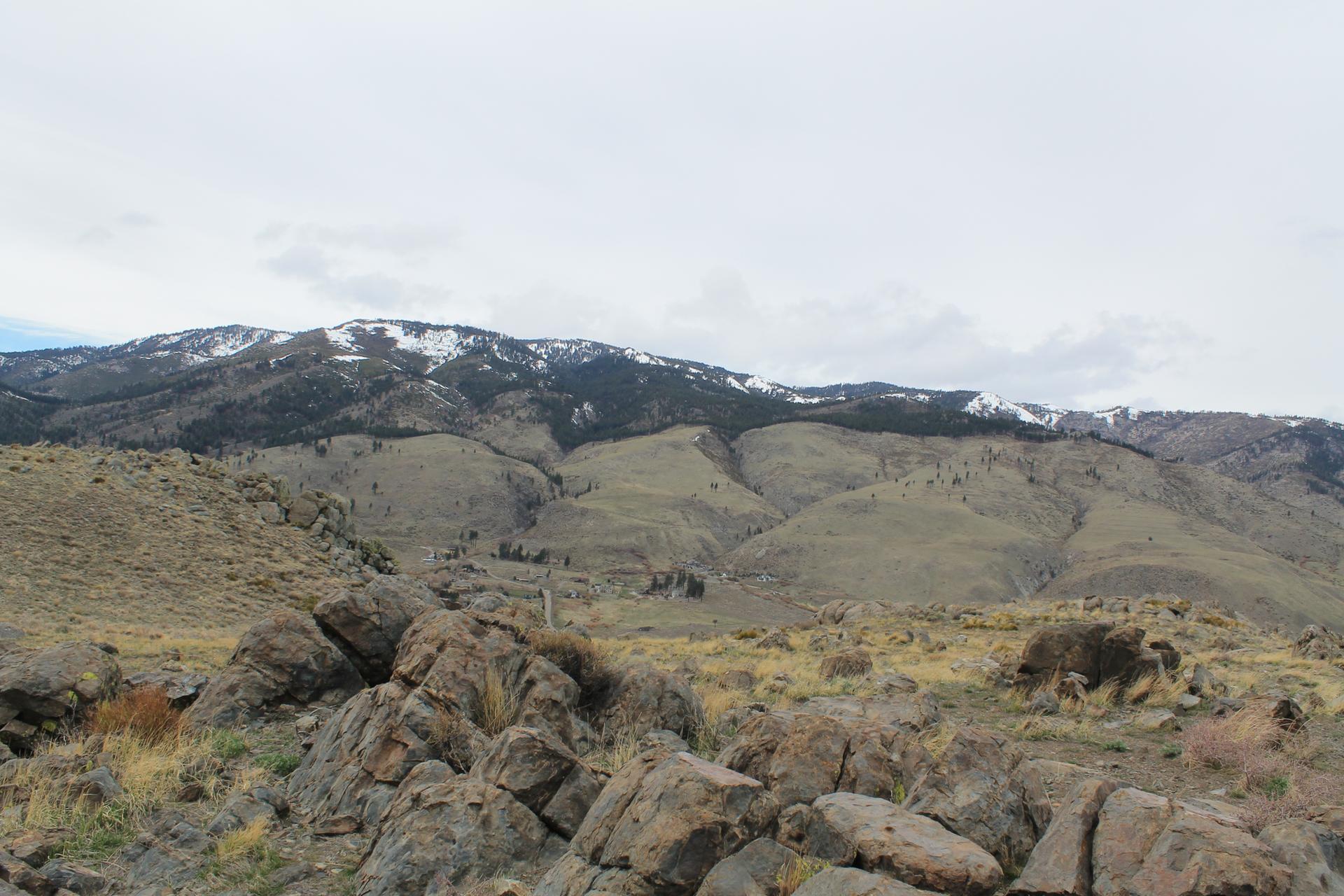 Rocky foreground with dry hills leading to snow-capped mountains under an overcast sky.