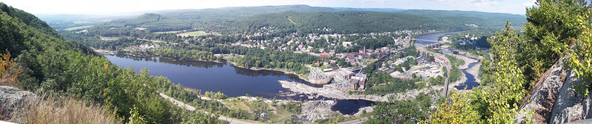 Panoramic view of a river, town, and forested hills under bright daylight.