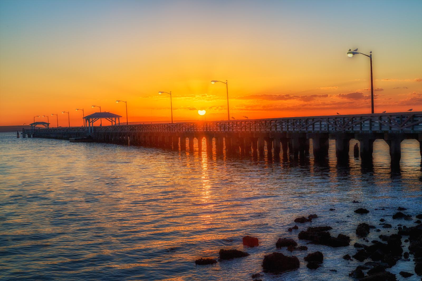 Long pier extending over water during a vibrant orange and yellow sunset.
