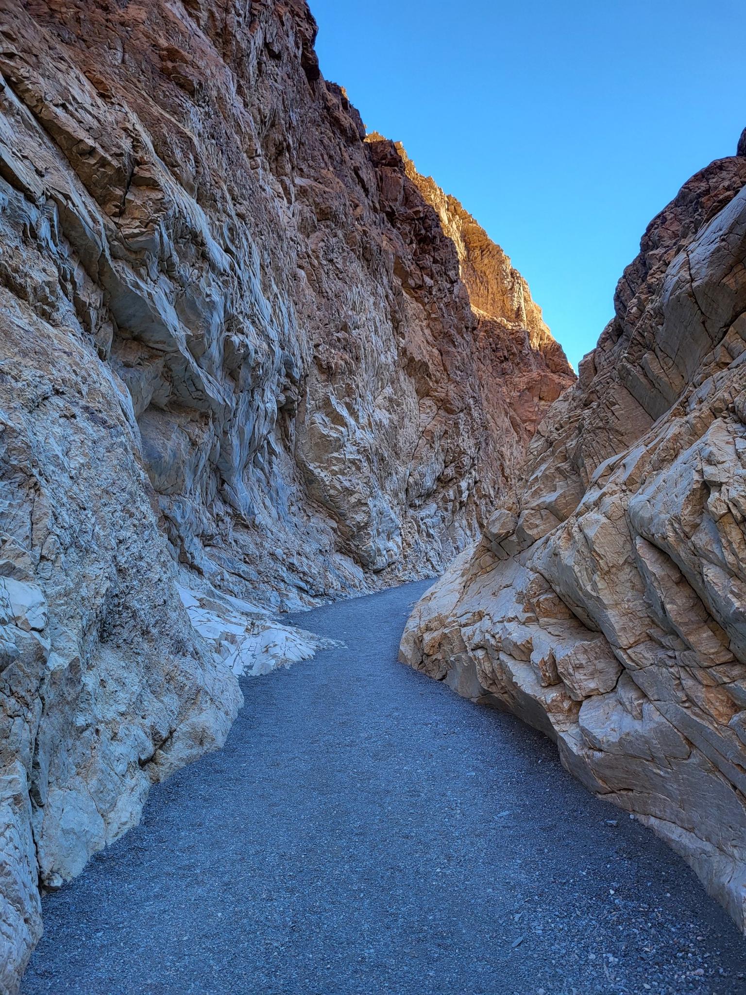 Narrow canyon path between steep, colorful rock walls in bright sunlight.