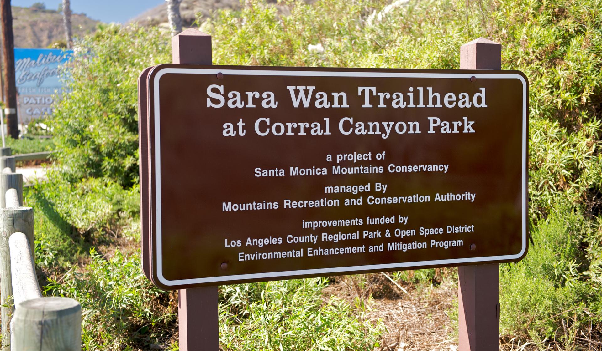 A brown trailhead sign with white text, surrounded by green bushes.