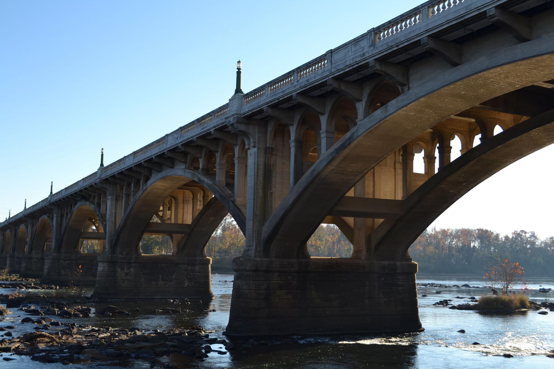 Ornate concrete arch bridge spanning a wide, rocky river under clear sky.
