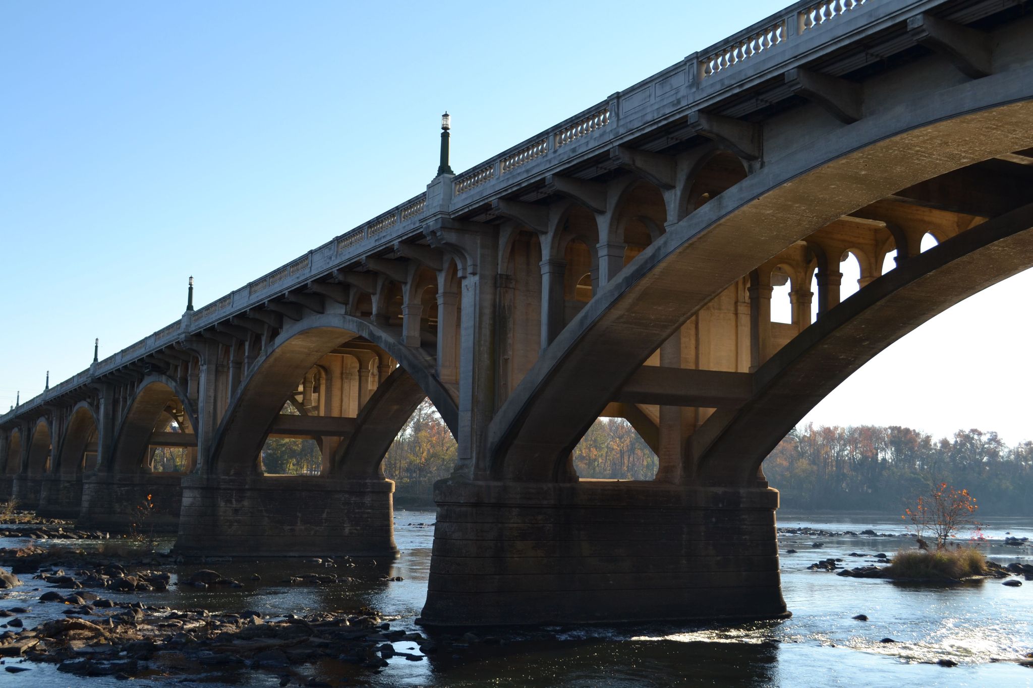 Ornate concrete arch bridge spanning a wide, rocky river under clear sky. - Three Rivers Greenway Trail