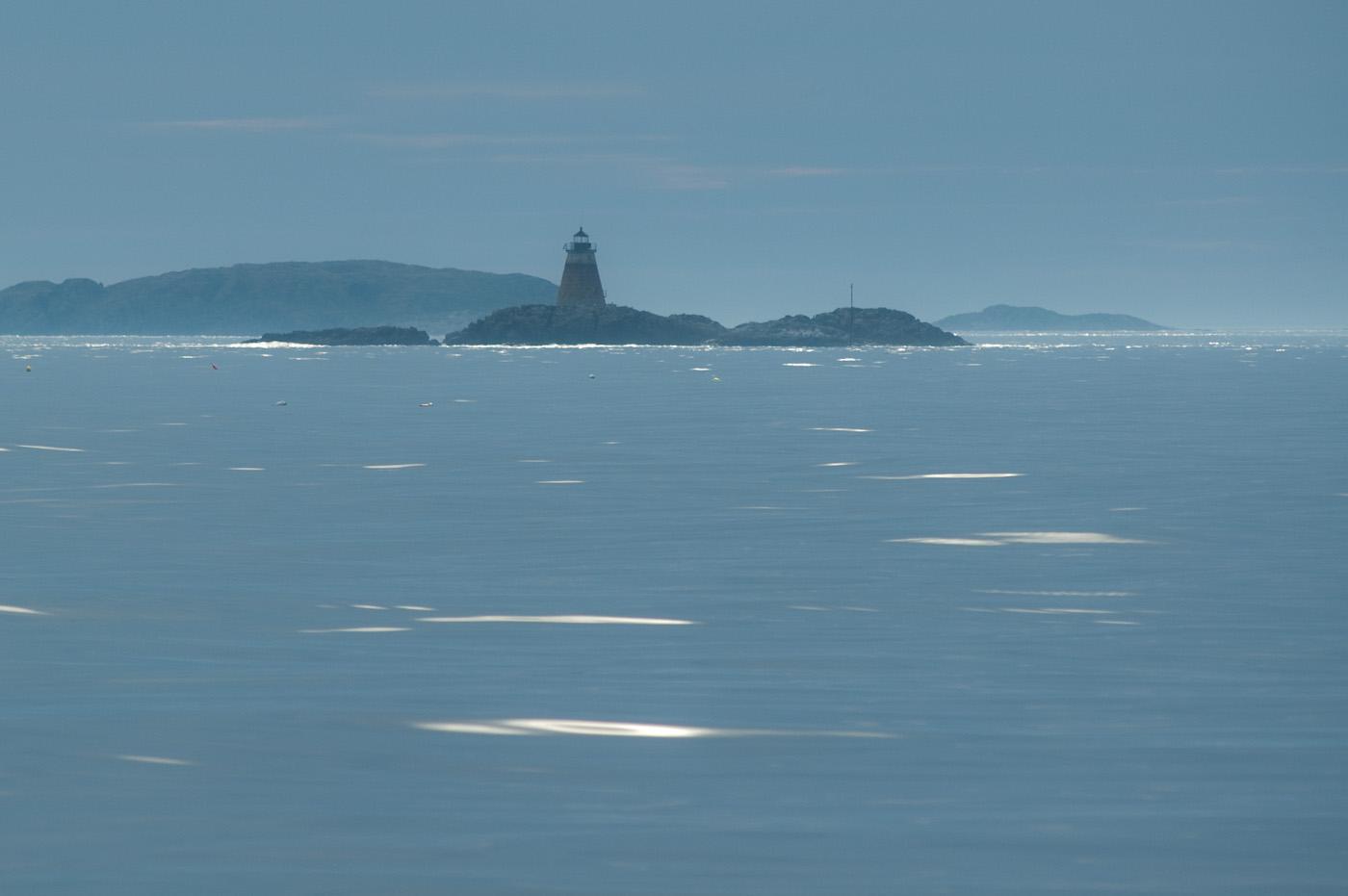 Distant lighthouse on a rocky island in a vast, hazy ocean.